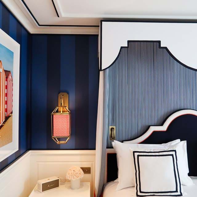 Detalhe de um quarto de luxo no AREV Hotel em Saint-Tropez, com uma elegante cabeceira azul, papel de parede listrado azul-marinho e um elegante arandela de parede.