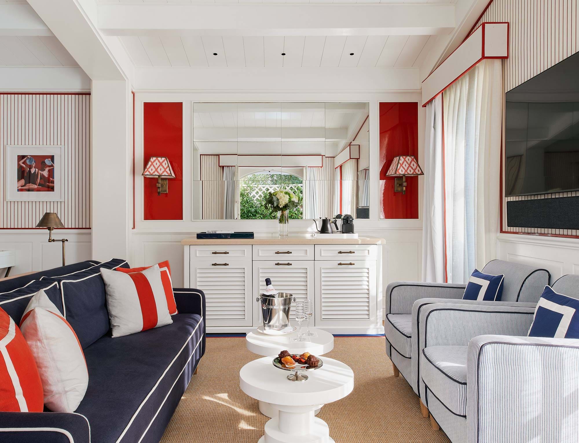 Área de estar da suíte chique com poltronas listradas, bar molhado branco e detalhes em vermelho no hotel AREV em Saint-Tropez.