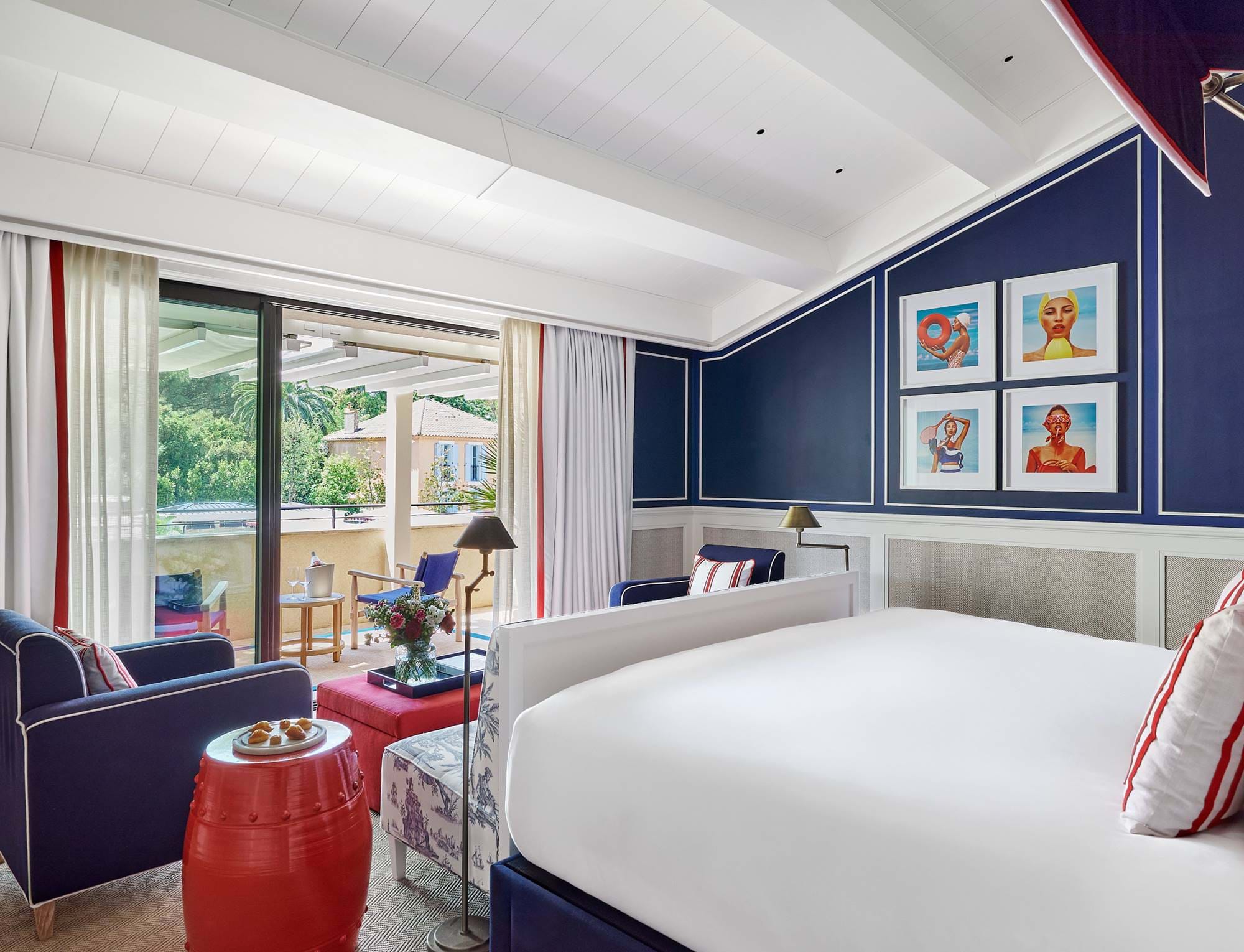 Um quarto luxuoso no hotel AREV em Saint-Tropez, com cama king-size, paredes azul-marinho com obras de arte modernas e um terraço privativo com vista para a vegetação exuberante.