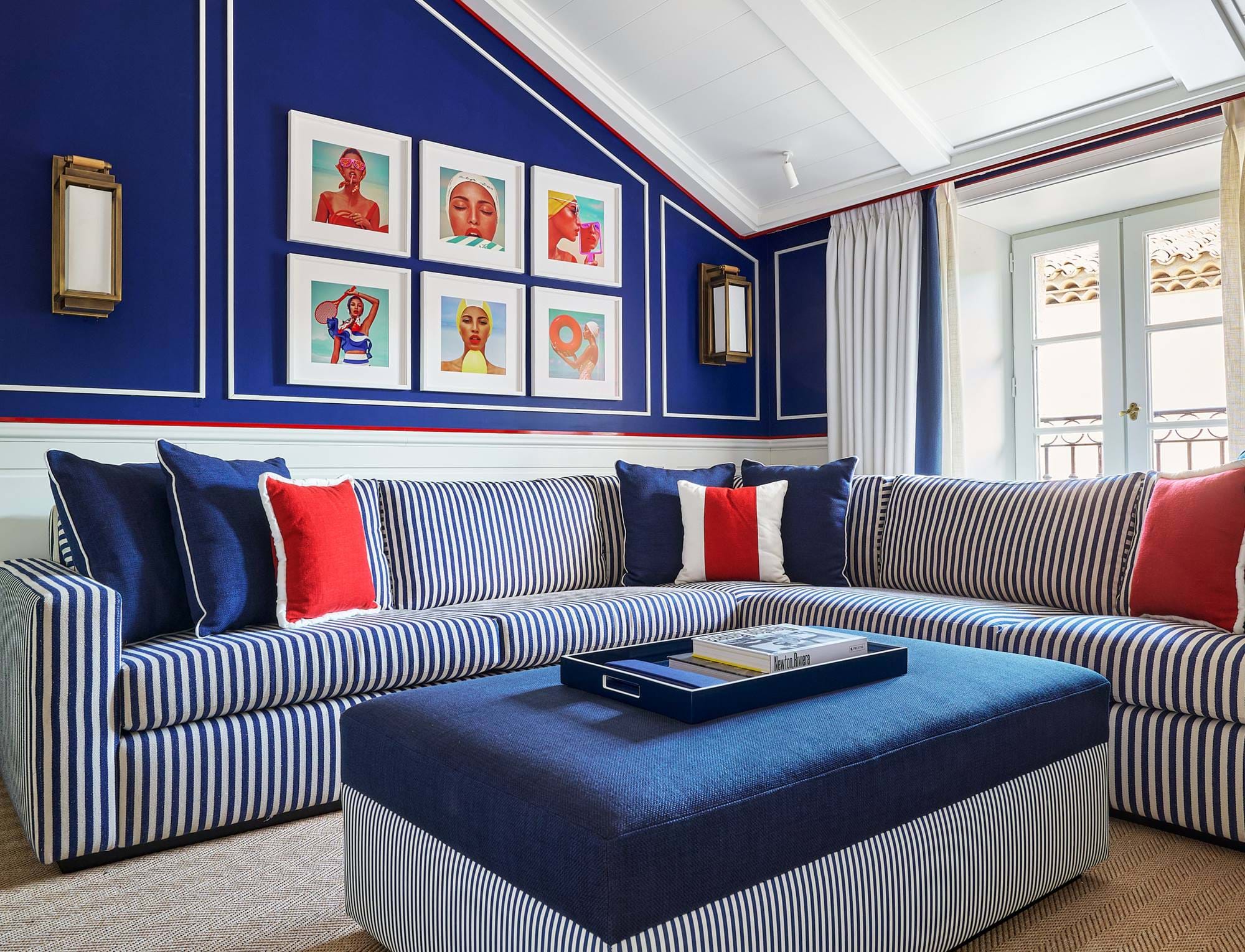 Suíte espaçosa no hotel AREV em Saint-Tropez, com grande sofá de canto cinza, parede azul intensa com quadros de arte e um pufe central.