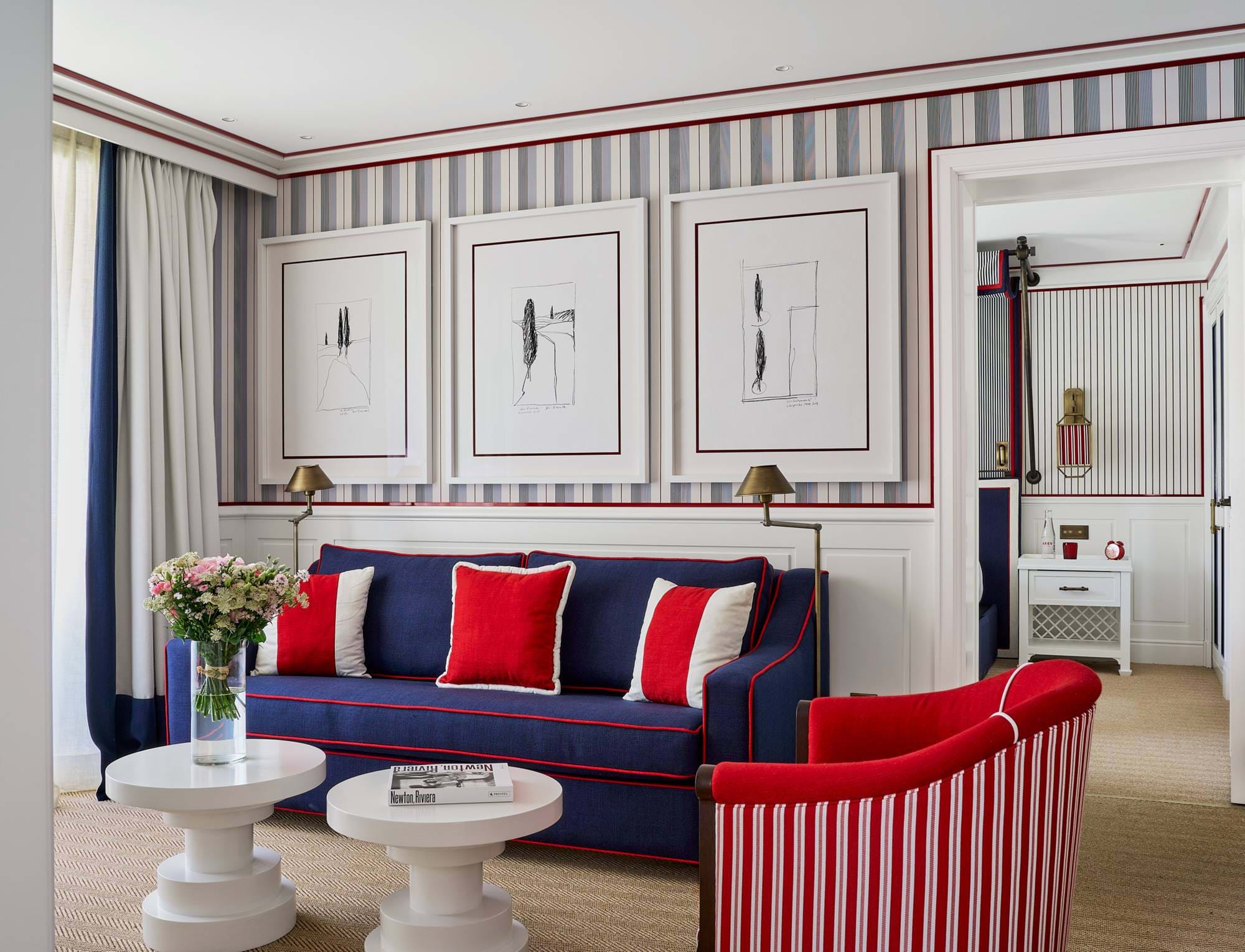 Área de estar chique no hotel AREV em Saint-Tropez, com sofá azul-marinho, almofadas vermelhas e brancas e papel de parede listrado sofisticado.