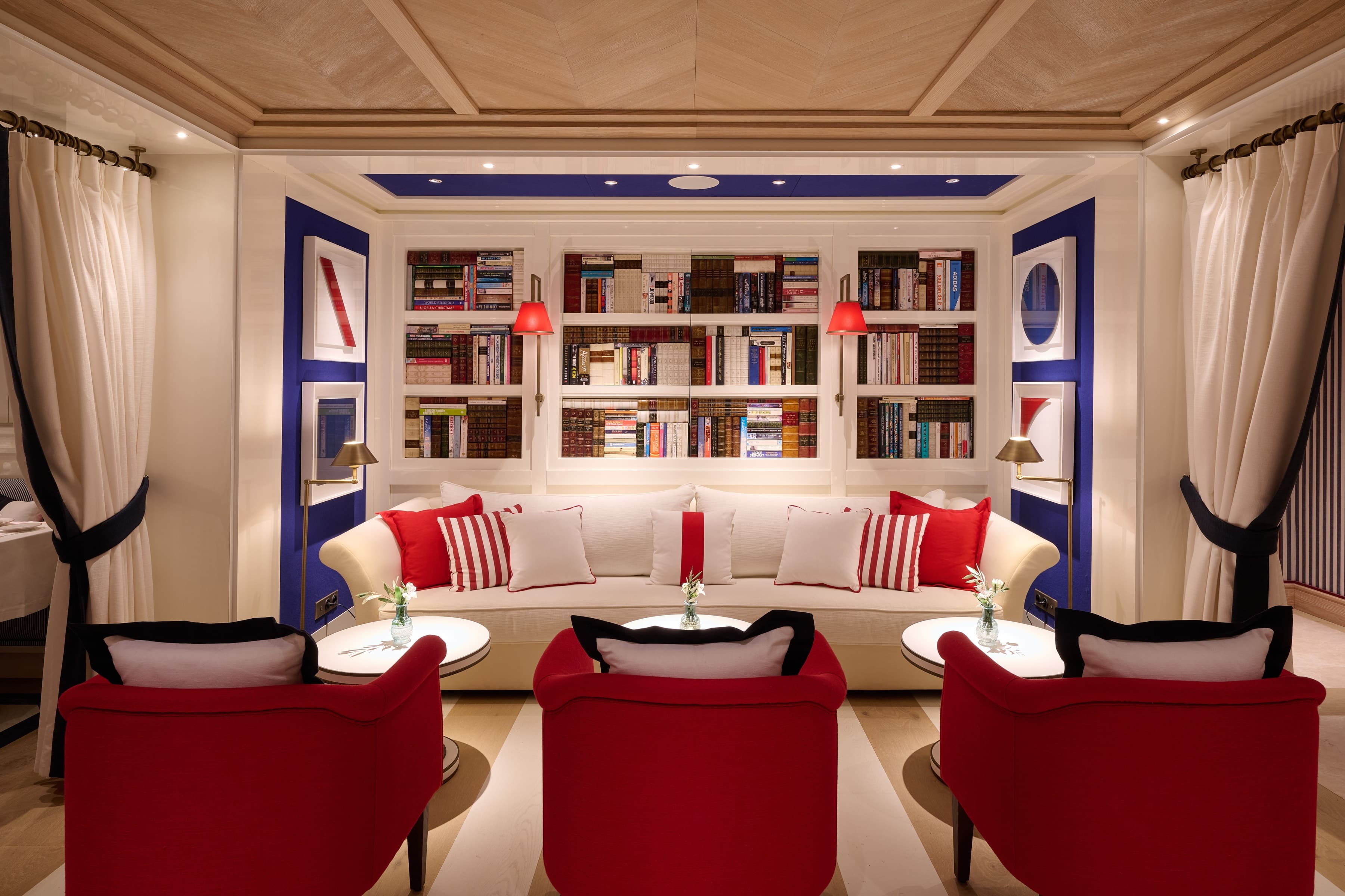 Biblioteca lounge do hotel Arev em Saint-Tropez, com grande estante branca, sofá creme e poltronas vermelhas num ambiente azul e branco