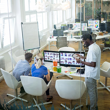 Three colleagues in an office collaborating on photo editing displayed on a computer monitor, with one standing and using a tablet.