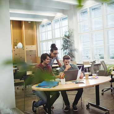 Three young professionals collaborating at a round table with laptops and documents in a bright modern office.