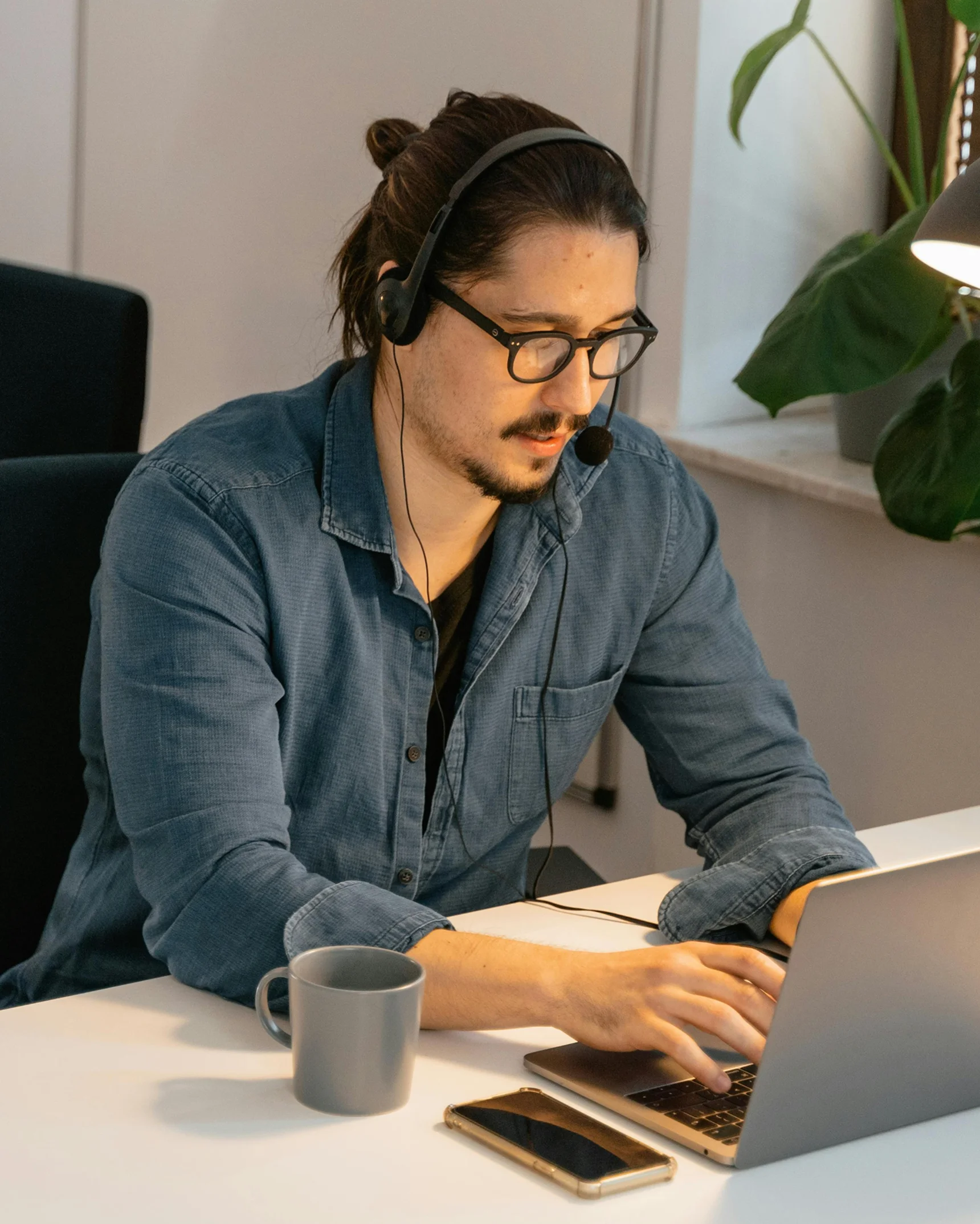Man with glasses and headset typing on a laptop at a desk with a gray mug and smartphone nearby.