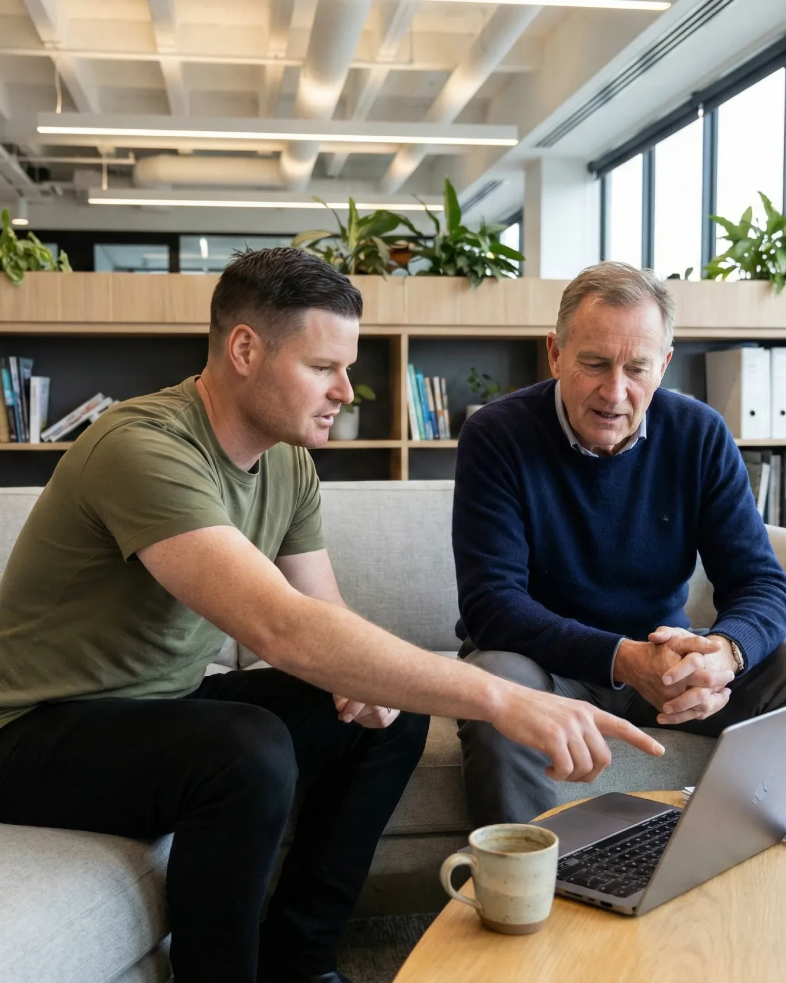 Two men seated on a couch looking at a laptop, with one man pointing at the screen, in a modern office setting.