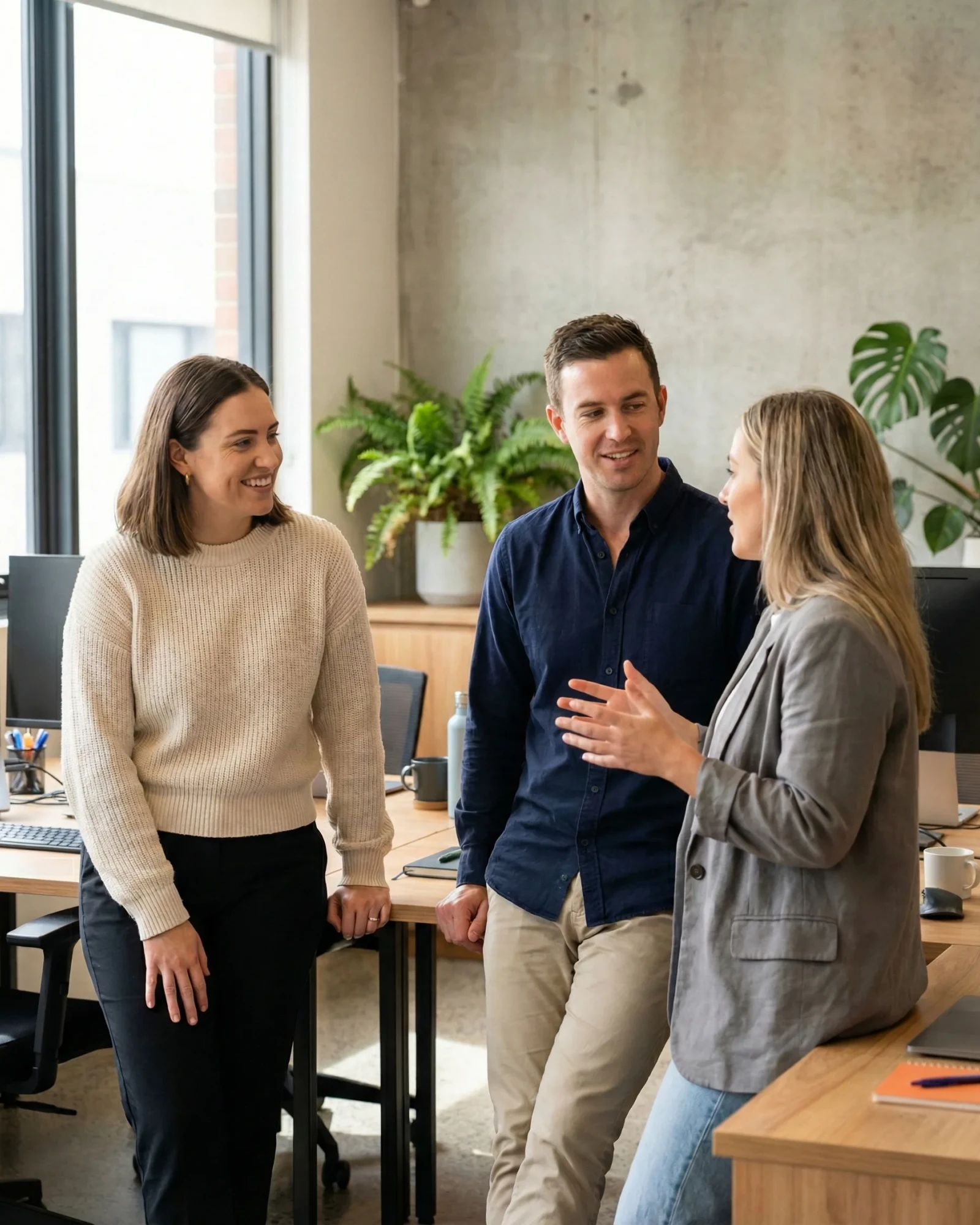 Three coworkers standing and having a friendly conversation in a modern office with plants and desks around.