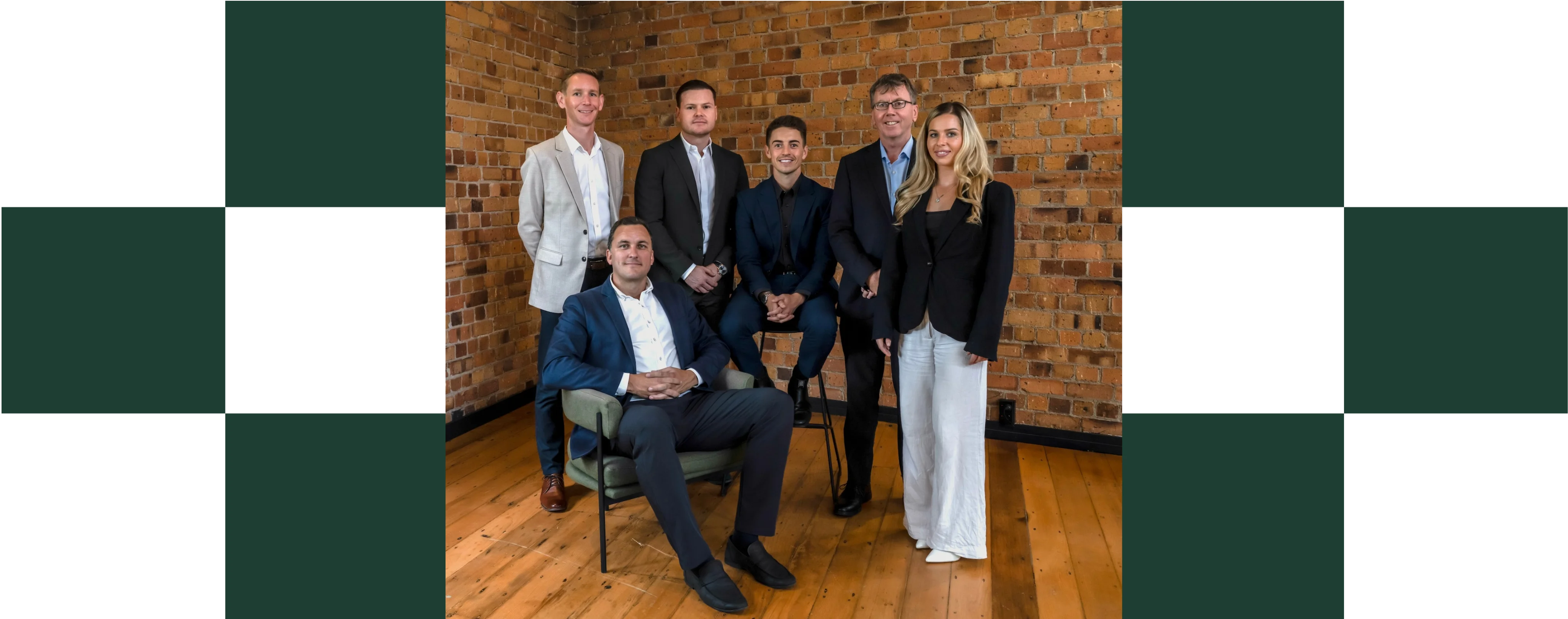 A professional team photo of six people posed against an exposed brick wall, with a man seated in a green armchair in front and five colleagues standing or seated on a stool behind him, all dressed in business attire (navy and beige suits, white blouse), set against a dark green and black geometric border frame.