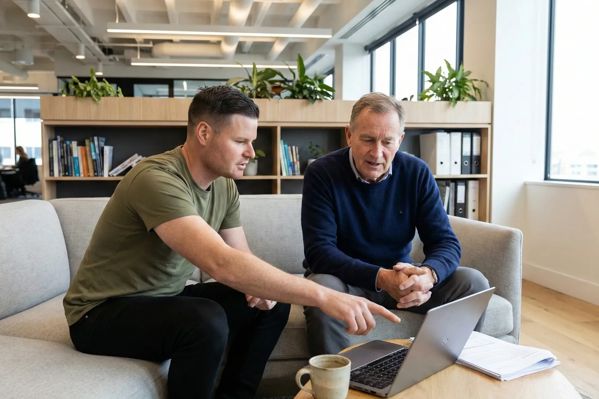 Two men sitting on a couch looking at a laptop with papers and a coffee cup on the table.