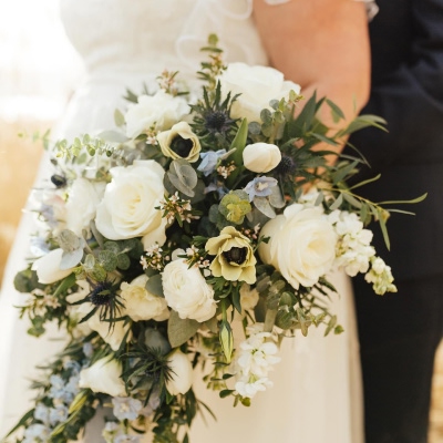 A cascading bridal bouquet featuring lush white roses, anemones, and delicate blue and green floral accents, complemented by eucalyptus and other soft greenery. The bouquet is held by a bride in a white gown, with the groom in a dark suit standing beside her.