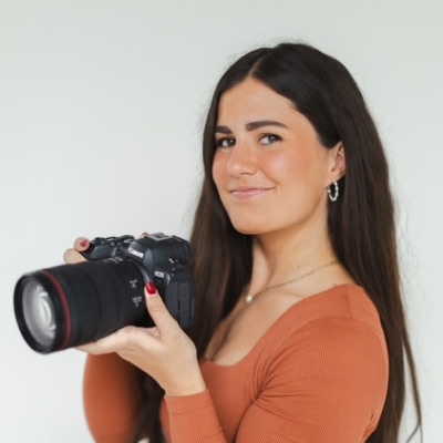 A young woman with long, dark brown hair and a warm smile holds a professional DSLR camera, looking confidently at the viewer. She wears a rust-colored top and silver hoop earrings.