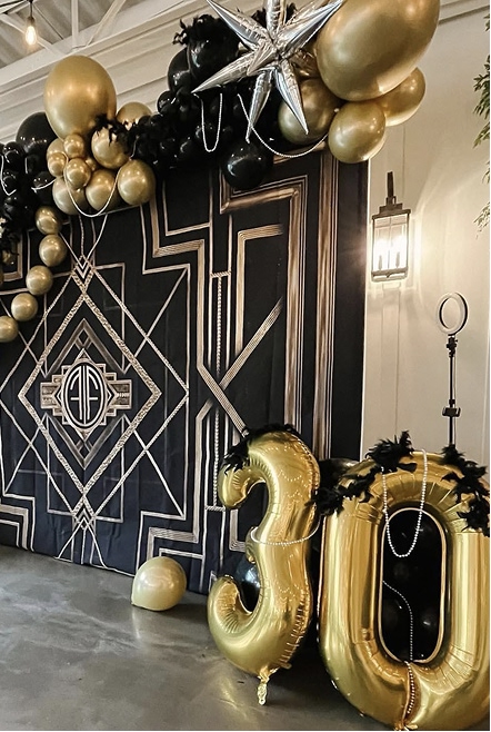 A black and gold Art Deco-themed event setup with balloon arrangements, a geometric backdrop, and large gold "30" balloons.