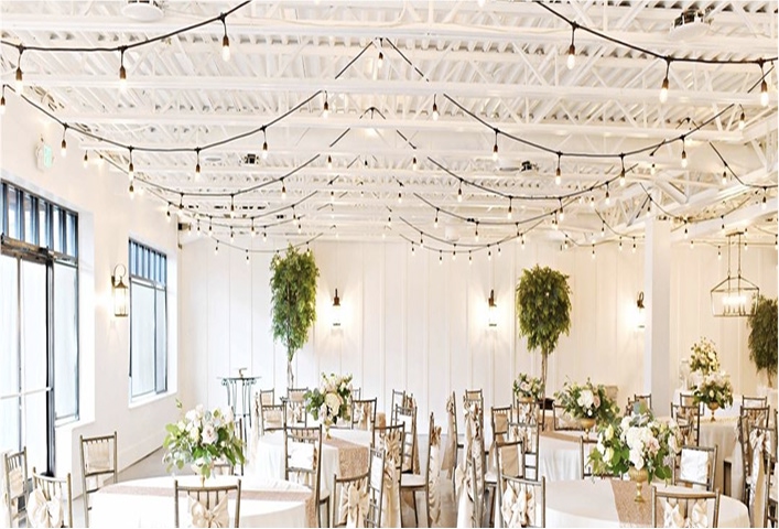 A beautifully decorated event space with white walls, bistro lighting, and round tables adorned with floral centerpieces.