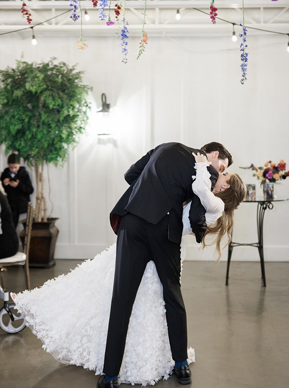 A groom dipping his bride for a kiss under hanging florals in a bright reception hall.