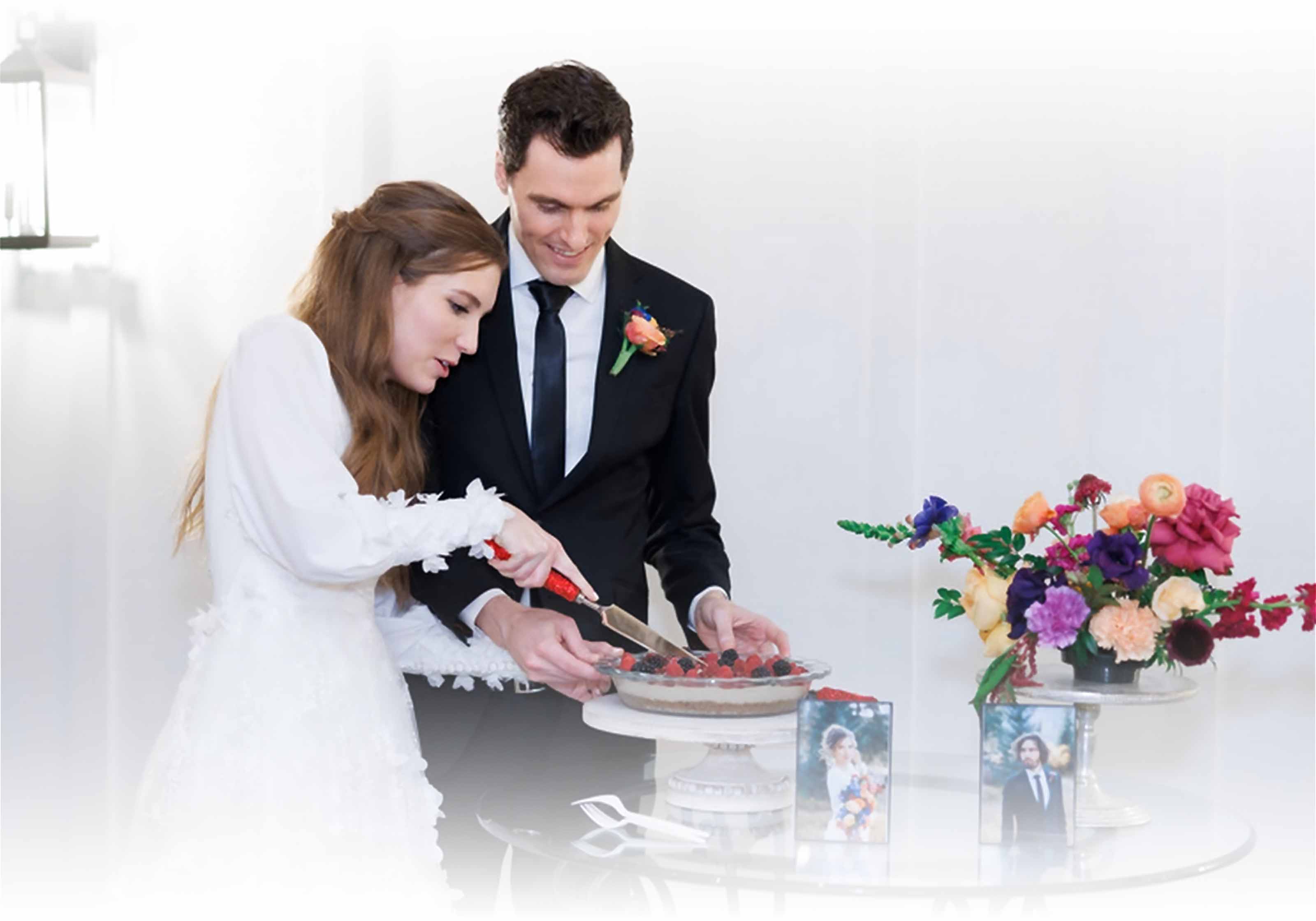Bride and groom cutting into a berry-topped wedding cake, surrounded by floral arrangements.
