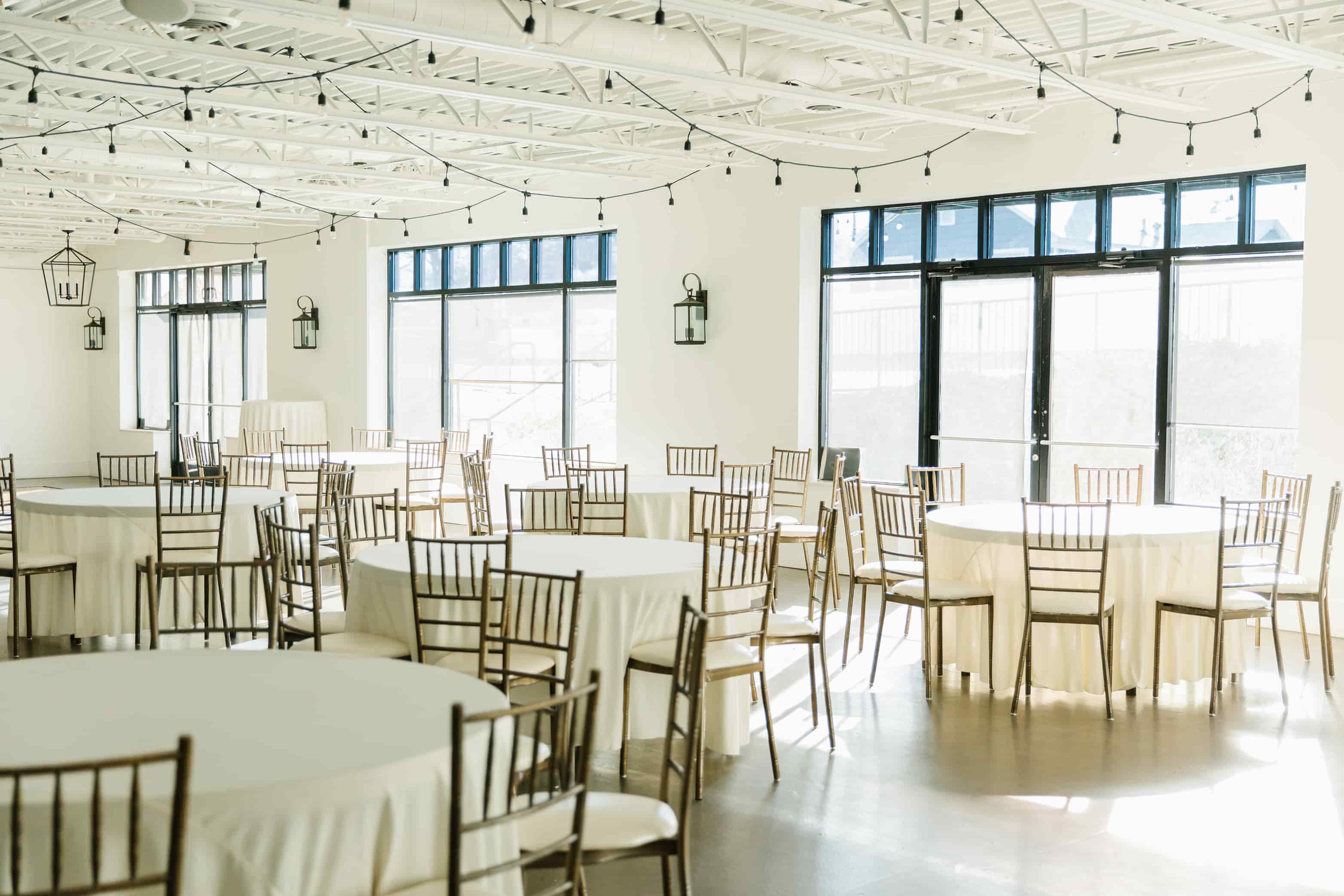Bright and airy event hall with round tables covered in white linens and surrounded by gold Chiavari chairs, set beneath exposed white ceiling beams and string lights, with large windows allowing natural light to flood the space.