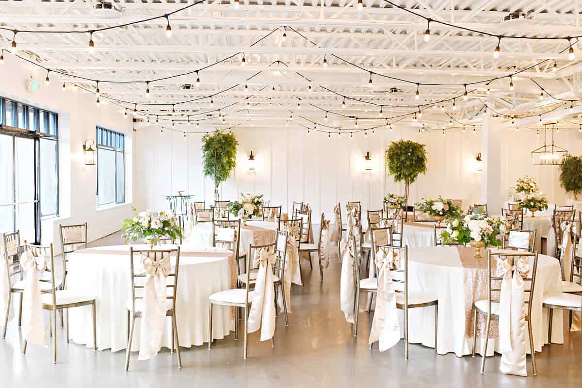 Elegant event hall decorated for a wedding or formal gathering, featuring round tables with white linens, gold chairs adorned with satin bows, floral centerpieces, overhead string lights, and potted trees against white paneled walls.
