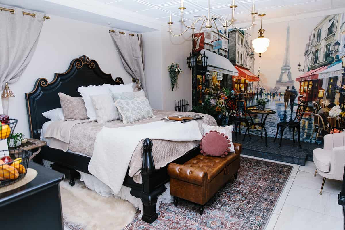 Cozy Paris-themed bedroom with a black ornate bed, soft neutral bedding, vintage rug, and a mural of a Paris street scene featuring the Eiffel Tower, café tables, and pedestrians in the background.