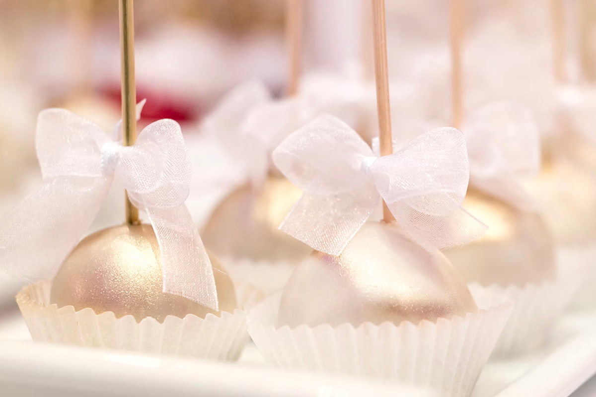 Gold-coated cake pops with white sheer ribbon bows on sticks, placed in white paper cups.