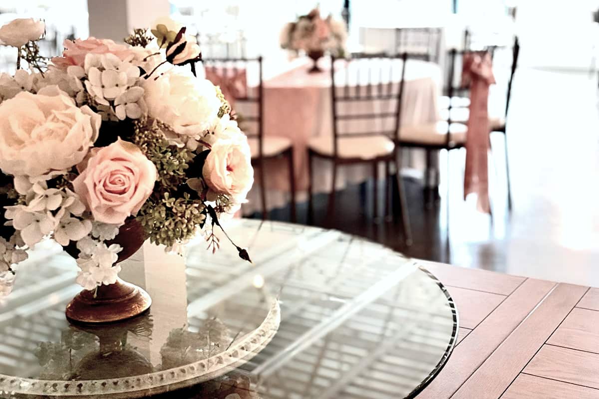 Close-up of a floral centerpiece with pink and white roses on a glass table, with a blurred background of a decorated event space with chairs and tables.