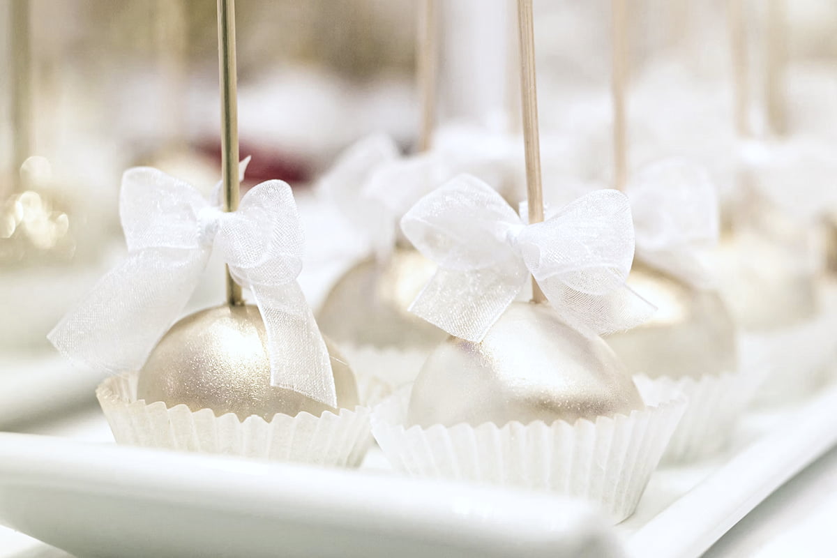 Gold-colored cake pops decorated with white sheer bows, arranged in white paper cups on a tray.