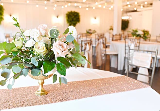 Elegant floral centerpiece with light pink and white roses and green foliage on a white table with a pink glitter runner in a softly lit reception hall.