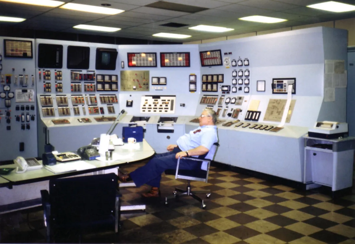 An operator seated at the console of a power plant control room, surrounded by panels of dials and switches