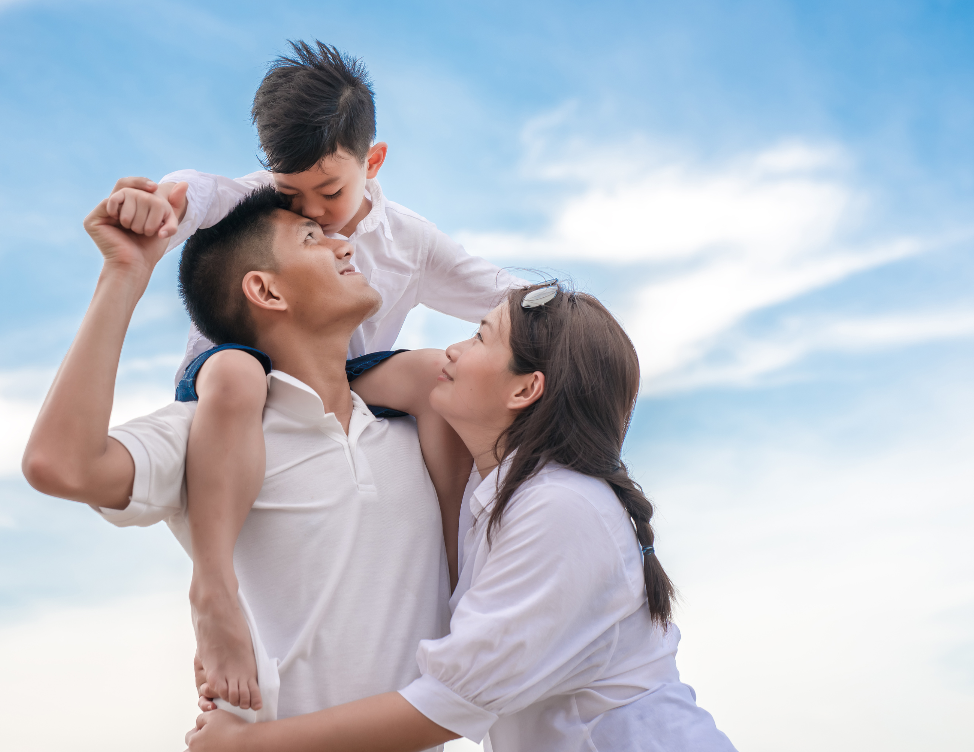 Man holding a boy on his shoulders while a woman embraces them both under a blue sky with clouds.