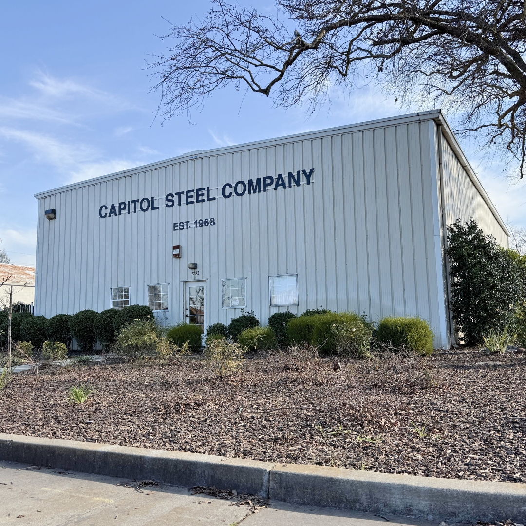 White metal building with the sign 'CAPITOL STEEL COMPANY EST. 1968' and bushes in front under a partly cloudy blue sky.