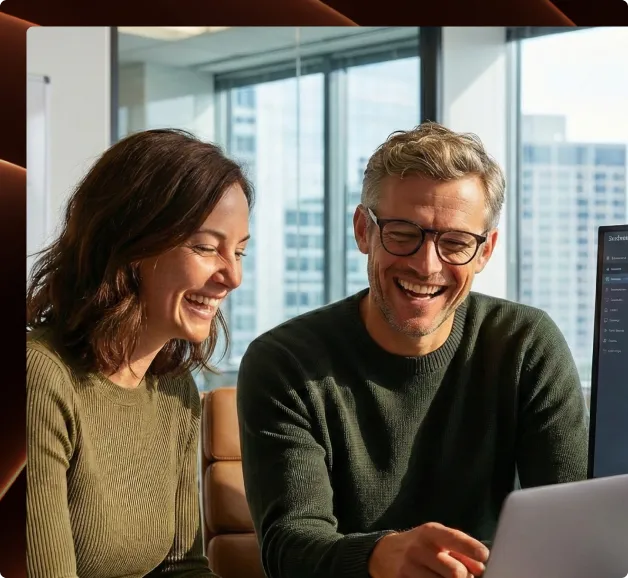 Smiling man and woman sitting indoors, looking at a laptop in a bright modern office with large windows.