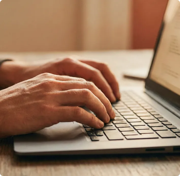 Hands typing on a laptop keyboard on a wooden table with a blurred screen in the background.