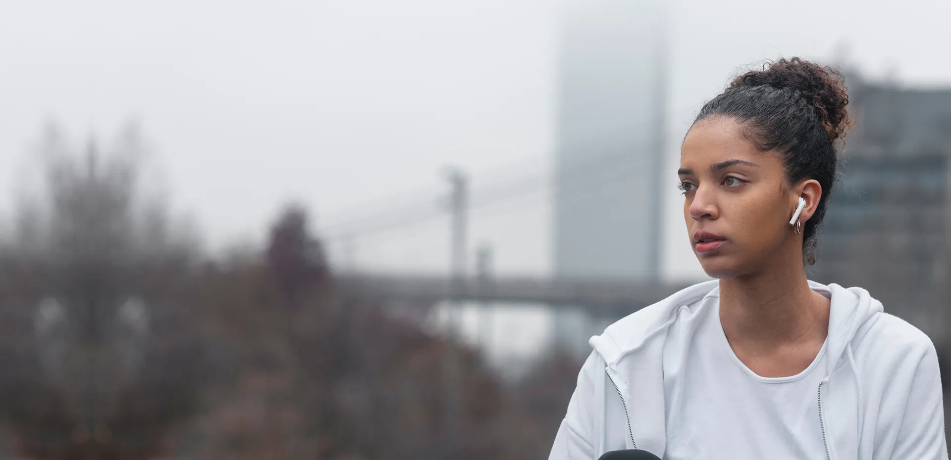 Young woman with curly hair in a bun wearing white earbuds and a white hoodie, looking to the right with a blurred bridge and trees in the background.