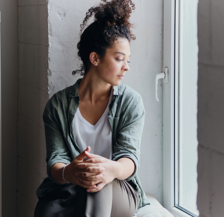 Young woman with curly hair tied up sitting by a window, looking outside thoughtfully.