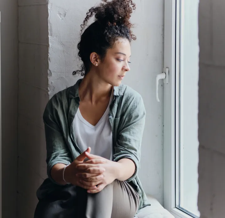 Young woman with curly hair tied up sitting by a window, looking outside thoughtfully.