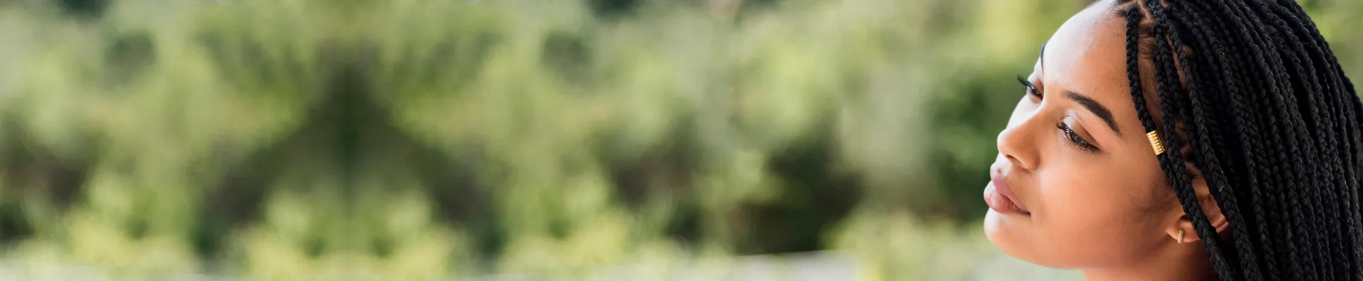Close-up profile of a woman with braided hair looking thoughtfully to the side against a blurred green outdoor background.