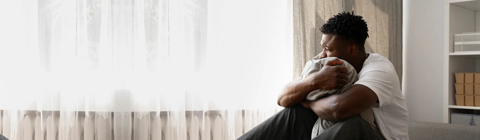 Pensive young man sitting on a couch near a window hugging a pillow and looking outside.