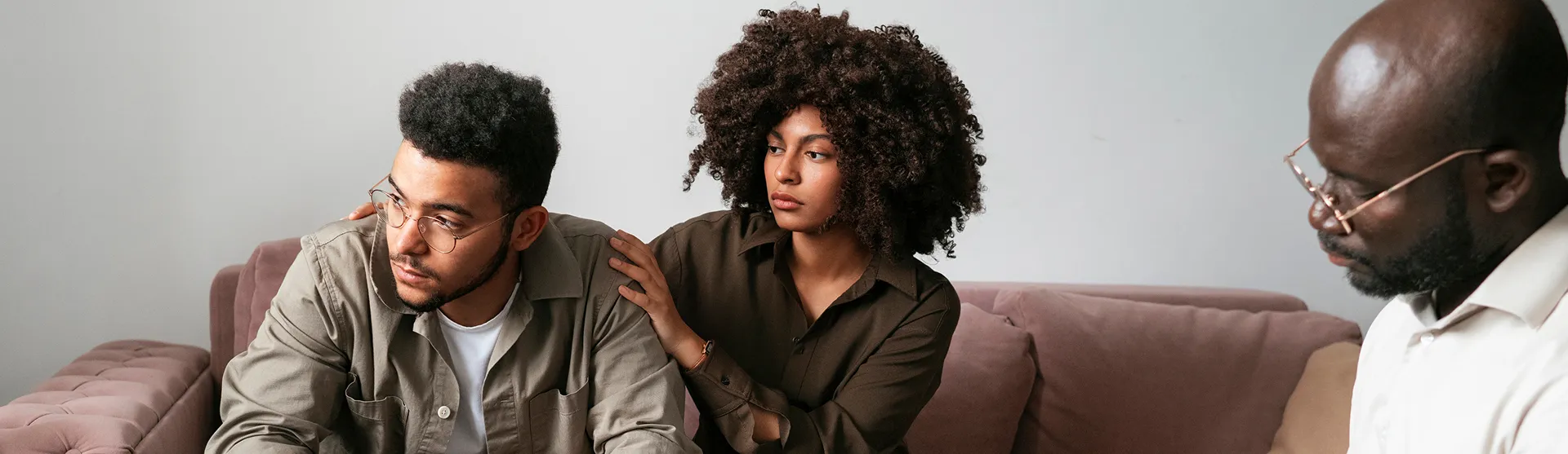 Woman comforting a man by placing her hand on his shoulder while a therapist listens during a counseling session on a couch.