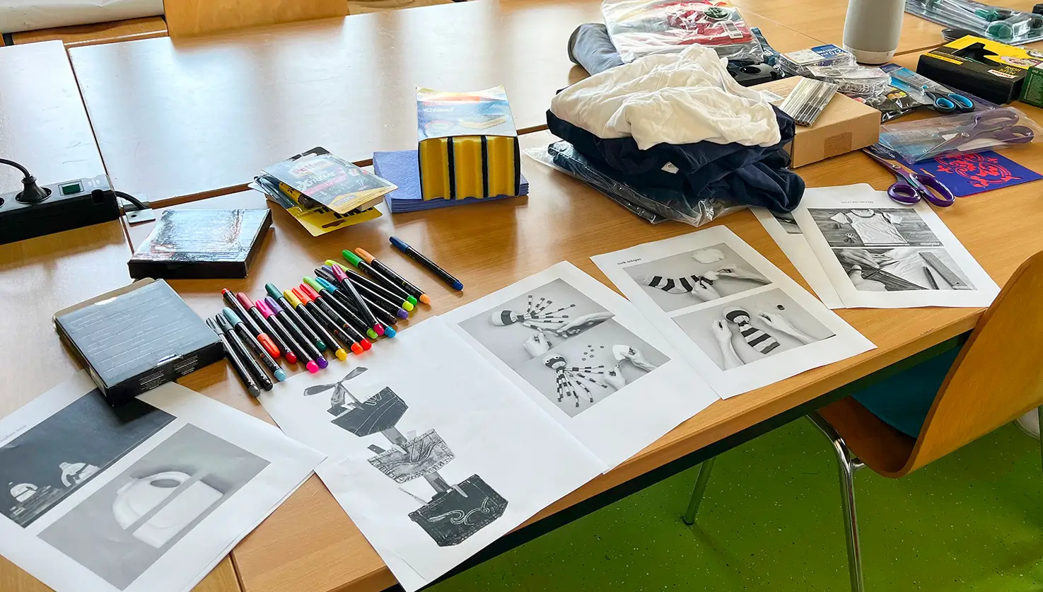 Auf einem Tisch liegen mehrere schwarz-weiße Drucke mit abstrakten und figürlichen Motiven, daneben liegen bunte Marker, Stoffe und weitere Bastelmaterialien.