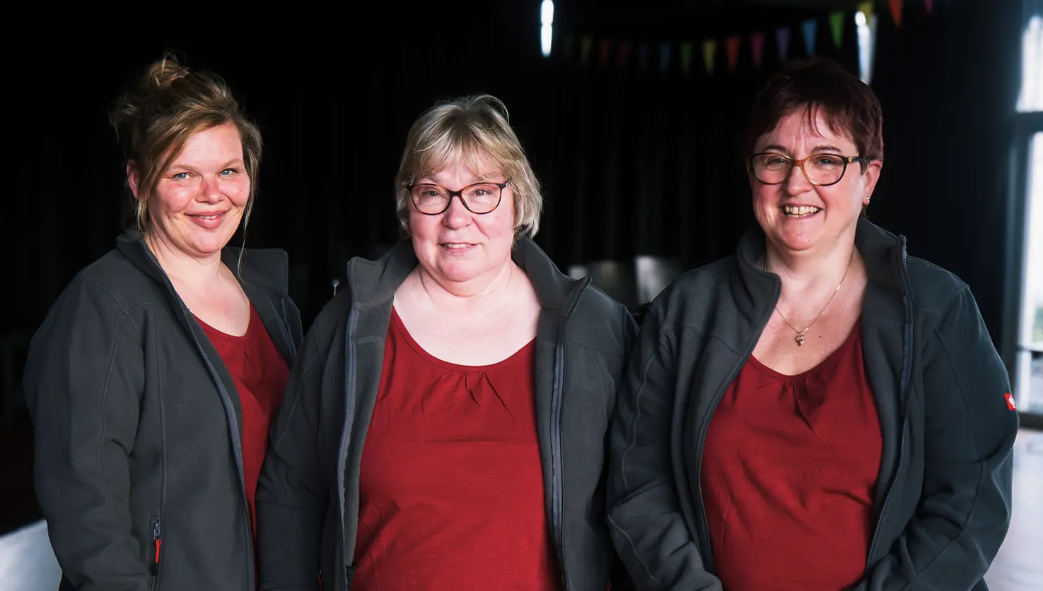 Drei Frauen in roten Oberteilen und schwarzen Jacken stehen nebeneinander vor dunklem Hintergrund.