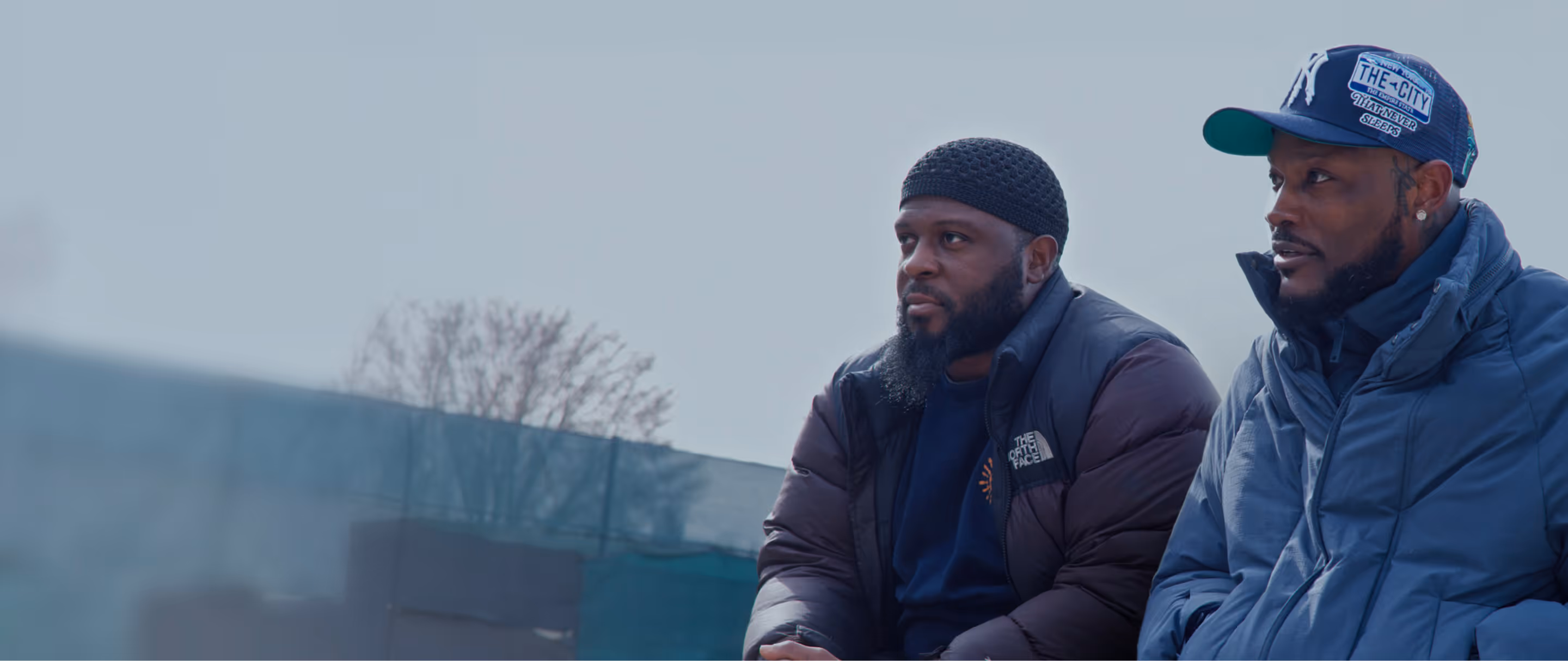 Two men in winter clothing sitting outdoors against a blurred background of trees and fencing.