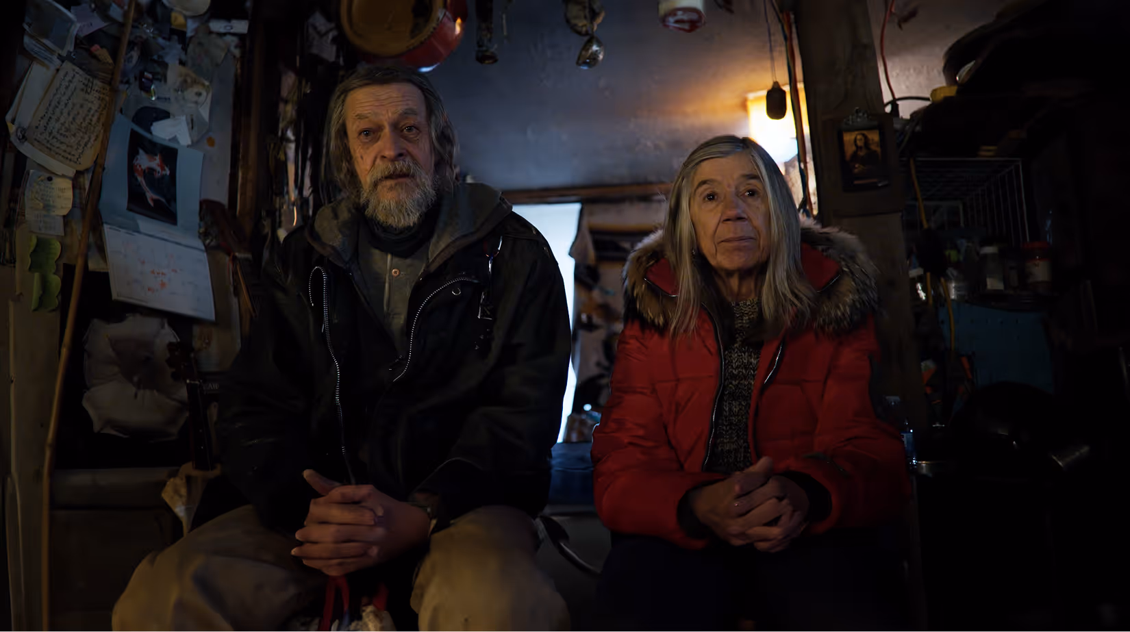 Elderly man with a beard and an elderly woman with gray hair sitting indoors in a dimly lit room.