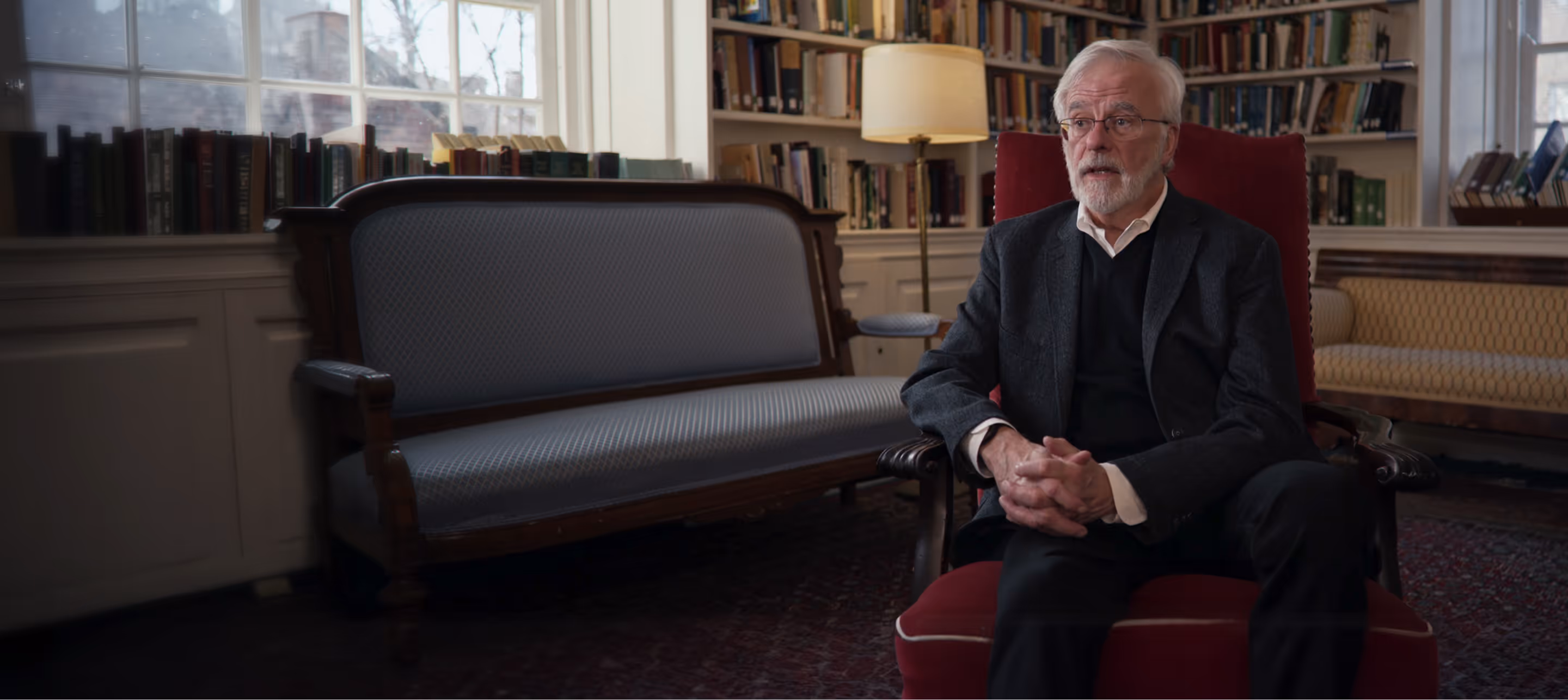 Elderly man with white hair and beard sitting and talking in a library room with bookshelves, sofas, and a standing lamp.