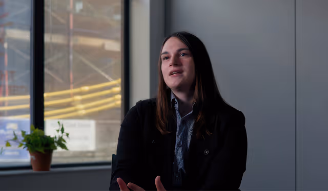 Man with glasses and floral shirt sitting cross-legged on a stool against an orange wall background.