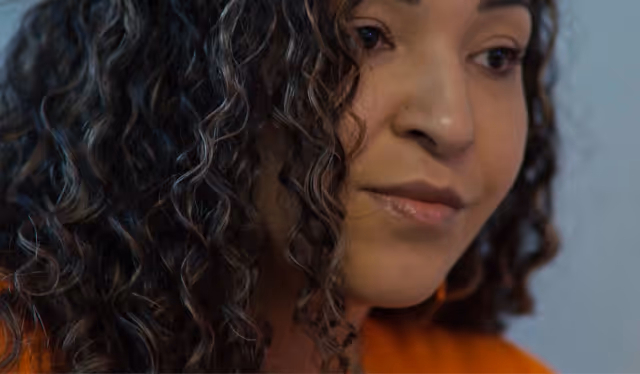 Person with long red hair wearing a red shirt with white hearts sitting at a desk working on a computer, with signs on the door saying 'Todos son bienvenidos aquí' and 'All are welcome here'.