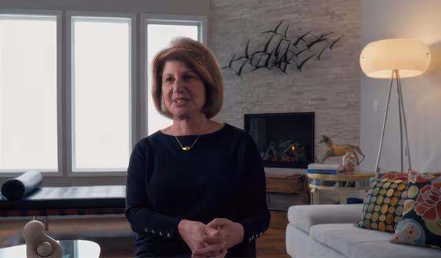 Elderly man with white hair, beard, and glasses speaking while seated in a red chair with a bookshelf and window in the background.