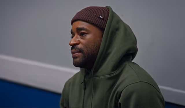 Man wearing a brown knit cap and green hoodie sitting against a gray and blue background.