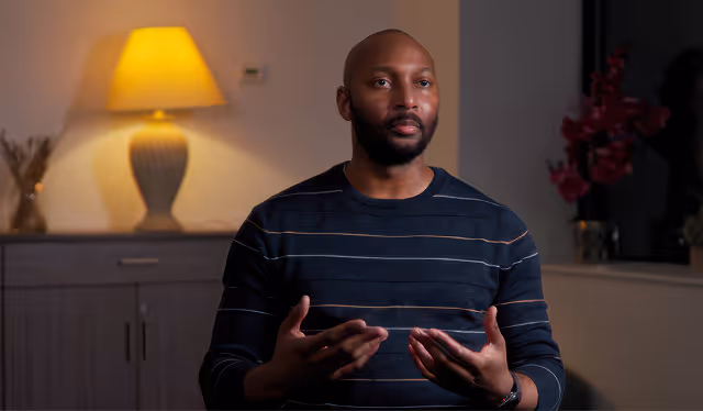 Man with a beard and striped sweater gesturing with his hands while seated indoors near a lit lamp and cabinet.