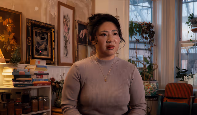 Woman in a beige sweater sitting in a cozy room with framed artwork, plants, and stacks of books behind her.