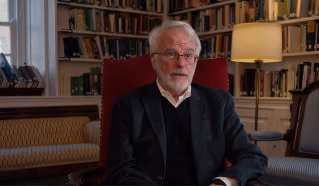Elderly man with glasses and gray hair sitting in a red chair in a room filled with bookshelves and a floor lamp.