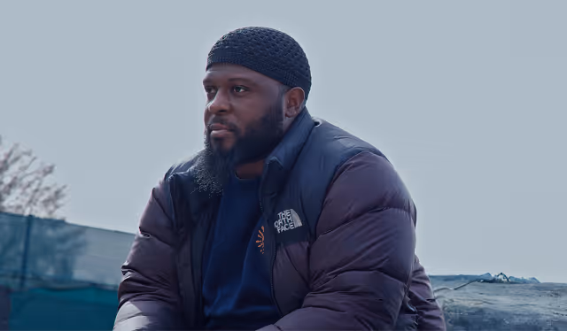 Man wearing a black knit cap and a puffy North Face jacket sitting outdoors with a neutral expression.